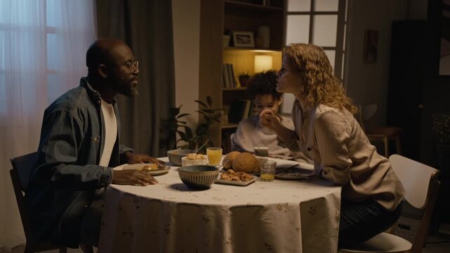 Side view shot of multiracial family conflict scene with parents fighting at dinner table, woman leaving room angrily with son watching
