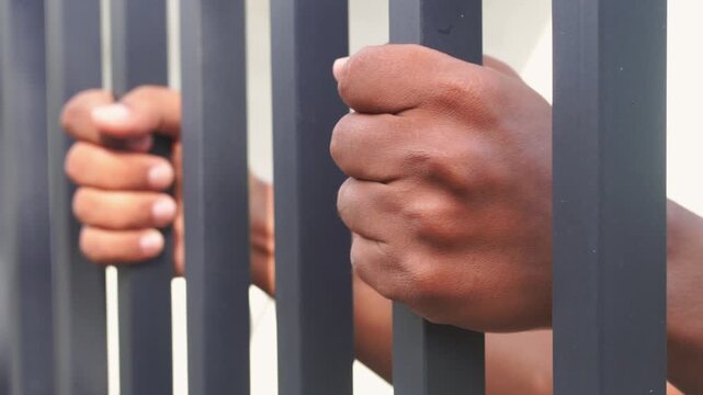 Close-up of hands gripping metal prison bars, symbolizing imprisonment, justice, crime, human rights, freedom restriction, and social issues. Concept of detention, punishment, and captivity.