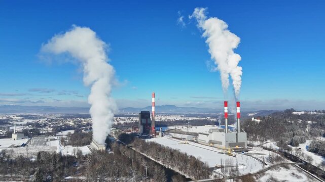 Aerial view of a power plant with tall chimneys emitting white smoke over a snowy landscape and distant mountains in Wildon, Styria, Austria.