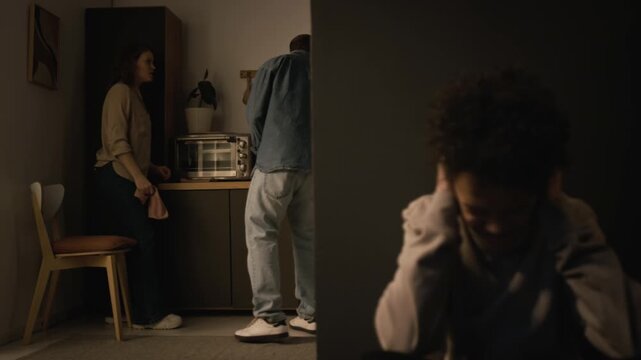 Family conflict scene with couple fighting and hitting each other in home kitchen with focus flowing to child hiding and covering ears in foreground, copy space