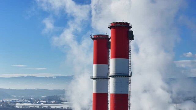 Aerial view of a tall red and white industrial chimney emitting thick white smoke against a clear blue sky and snowy landscape in Wildon, Styria, Austria.