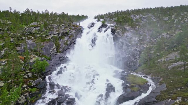 Aerial view of Kjemafossen waterfall cascading over dark rocks surrounded by lush green pine forests under a cloudy sky in Saltdal, Nordland, Norway.
