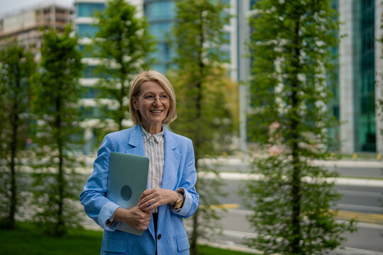 Mature businesswoman holding laptop walking in city park