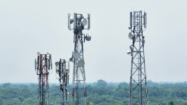 Aerial view of several telecommunication towers with various antennas and satellite dishes standing above a dense green forest under a clear sky.