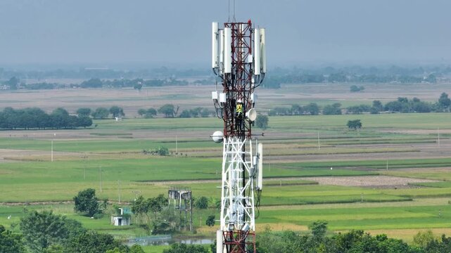 Aerial view of a red and white telecommunications tower with a technician in a yellow vest performing maintenance over vast green agricultural fields and rural landscape.