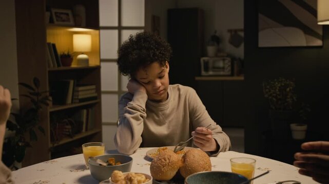 Zoom out shot of upset Black teenage boy sitting at dinner table at home and picking food while watching parents fight and yell at each other