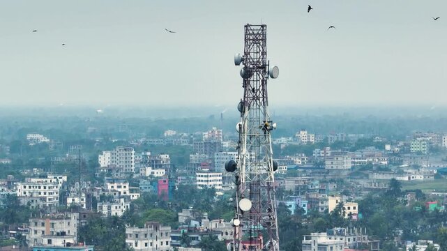 Aerial view of a tall telecommunication tower with antennas and satellite dishes overlooking a dense urban landscape with residential buildings Bangladesh.