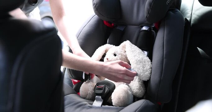 Danger prevention. Woman unfastening toy bunny in child safety seat inside car, closeup