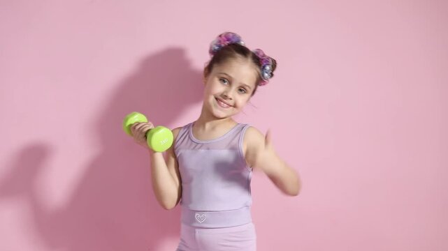 Girl exercising with dumbbell and showing thumbs up on pink background. Child's sports