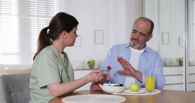 Caregiver with breakfast and elderly man refusing to eat it at wooden table indoors