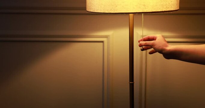 Woman turning on lamp with pull-chain switch near light wall at home, closeup