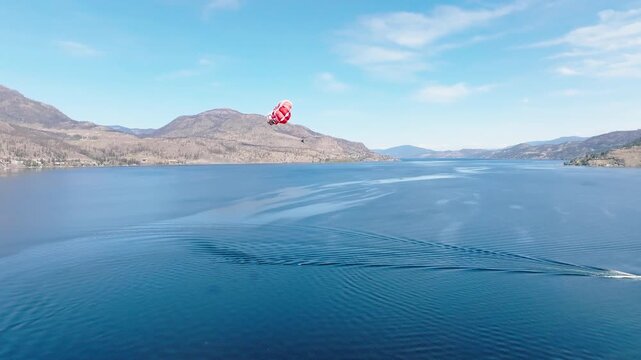 Parasail with the design of a Canadian maple leaf flag, high above a lake with desert mountain in the background. A boat can be seen towing the parasailer. 