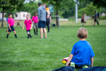 Fototapeta premium Children play soccer in a park while one boy watches from the grass in a warm afternoon setting