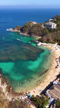 Vertical aerial view of a crowded tropical bay with turquoise water full of coral reefs, beach palapas and a clear sky, Huatulco Oaxaca Mexico, 4K 60fs flyover tilt-down drone shot