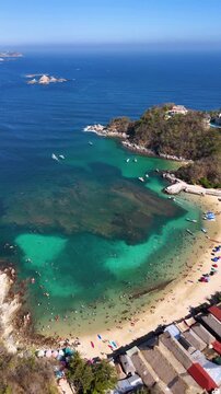 Panoramic vertical aerial view of a crowded tropical bay with turquoise water full of coral reefs, beach palapas and a clear sky, Huatulco Oaxaca Mexico, 4K 60fs orbit arc drone shot