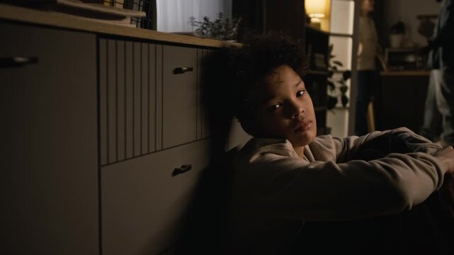 Side view shot of upset Black teenage boy sitting on floor in dark room and turning looking at camera with parents fighting in blurred background, copy space