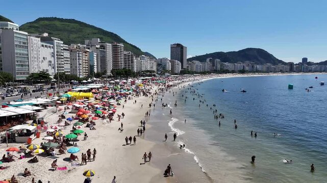 Aerial drone footage traveling parallel to Copacabana Beach, showcasing the curved shoreline, beach activity, ocean waters, waterfront skyline, coastal mountains and vibrant urban landscape in Rio de 