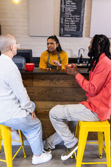 Indian barista in mustard top speaking, gesturing behind cafe bar while customers holding red mugs © wavebreak3