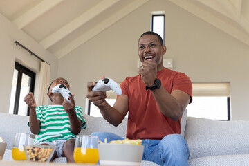 African American father and son cheering, holding white controllers on sofa with popcorn and juice © wavebreak3