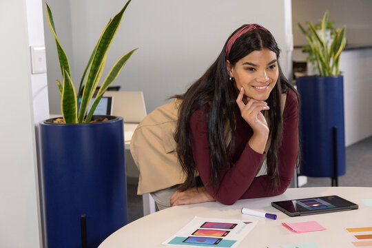 Mid-adult woman wearing maroon top leaning over round table in office, reviewing tablet mockups