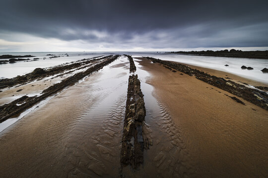 View of the unique flysch rock formations on the sandy shore of Playa de Sopelana under a dramatic cloudy sky in Vizcaya, Pais Vasco, Spain.