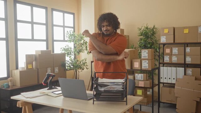 Man with package at office desk, arms raised in a celebratory stretch amid stacked cardboard boxes and laptop in a building; small business success joy.