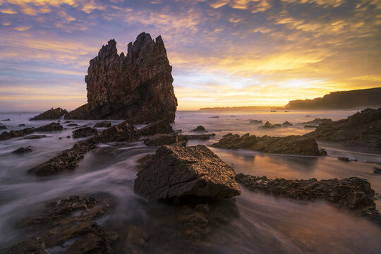 View of the iconic sea stack at Playa de Portizuelo with misty water and a golden sunset sky over the rocky coastline Spain.