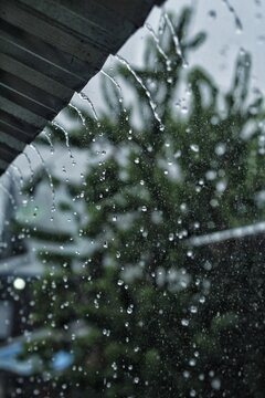 Close-up of heavy rain pouring down from a metal roof against a blurred background. Water pouring from a rooftop with a moody, dark green nature background.