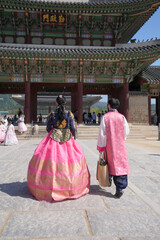 Fototapeta premium Two people walk toward a traditional Korean gate. One wears a vibrant pink hanbok with gold embroidery. The other carries a woven bag, dressed in matching pink robes