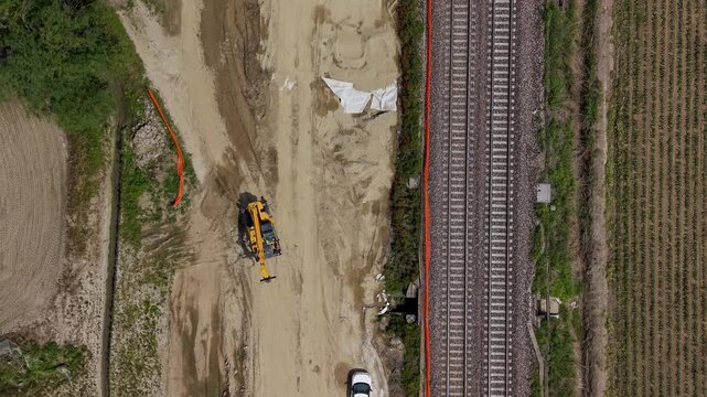 Railroad Construction Site Aerial View: An aerial perspective of a railroad construction site, showing an excavator at work amidst sand and dirt, with railway tracks running parallel to a rural field.