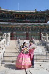 Naklejka premium Couple stands before a historic Korean palace. They wear traditional hanbok with vibrant patterns. Their smiles show joy in this cultural setting. The bright sky enhances the grand architecture