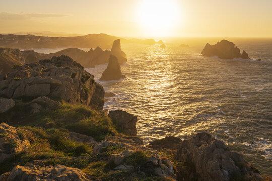 View of the rugged coastline and sea stacks under a golden sunset with waves crashing against rocky cliffs in Liencres, Cantabria, Spain.