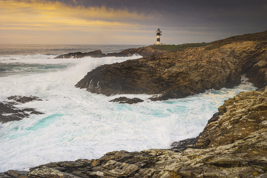 View of Illa Pancha lighthouse standing on rugged cliffs as powerful waves crash against the rocks under a golden sunset sky Lugo, Lugo, Spain.