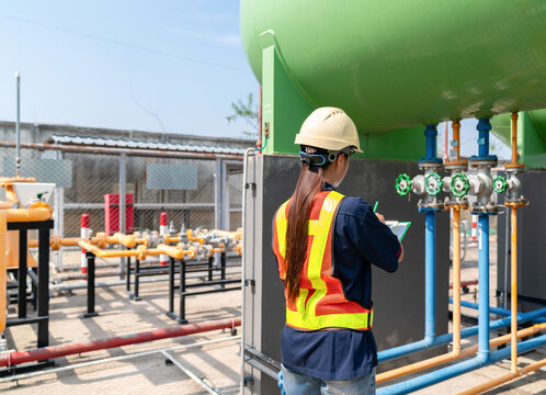 Rear view of a female safety officer or engineer in a hard hat and reflective vest performing an inspection of large industrial storage tanks and pipeline valves.