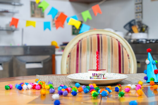 Festive birthday party table with happy birthday plate, colorful pompom, flag bunting and wicker chair in decorated kitchen interior, celebration concept.