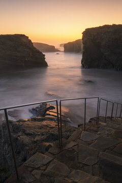 View of Praia das Catedrais with stone steps, metal railings, and dramatic rock formations during a golden sunrise over misty waters in Lugo, Lugo, Spain.