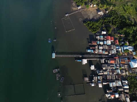 Aerial view of Galang Baru coastal village with houses on stilts, a long pier, and floating fish farms along the turquoise shoreline in Galang, Riau Islands, Indonesia.
