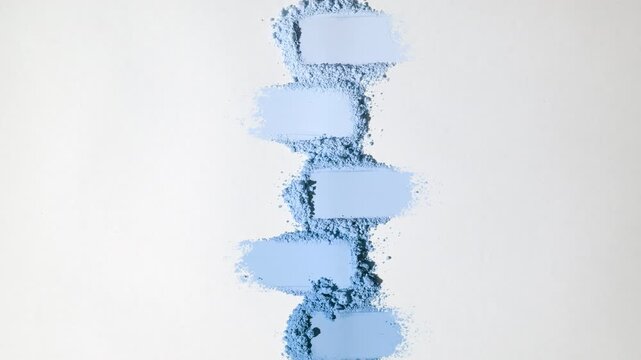 Top-down vertical shot of five blue organized powder smears on a faint grey backdrop highlighted by rotating directional light producing ever-changing shadows
