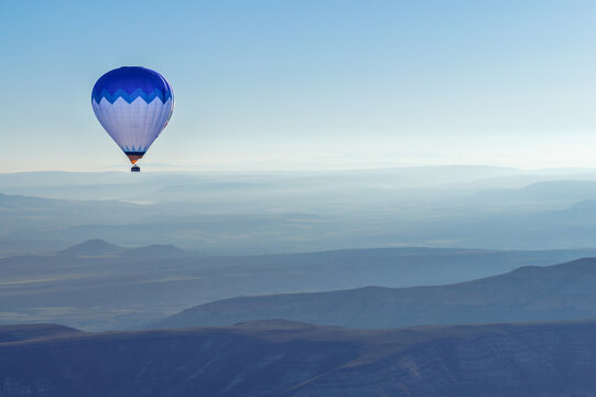 Aerial view of a blue and white hot air balloon floating over the misty volcanic landscape and layered ridges of Cappadocia in Kayseri, Kayseri, Turkiye.
