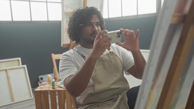 Man painter in studio holds smartphone and scrolls with thumb while seated by easel and canvases, wearing apron and checking reference images; creative focus.