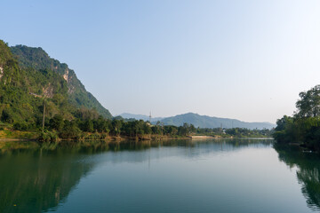 Fototapeta premium Calm river reflects distant hills and lush greenery. Sloping mountain covered in dense vegetation. Sky clear and blue, with soft haze on horizon. Trees line riverbanks, offering natural framing