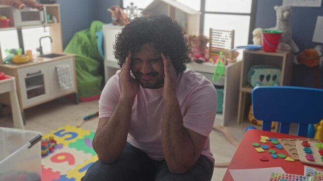 Man rubbing forehead and holding head in kindergarten building among toys and small table, foam number mat visible; caregiver fatigue.