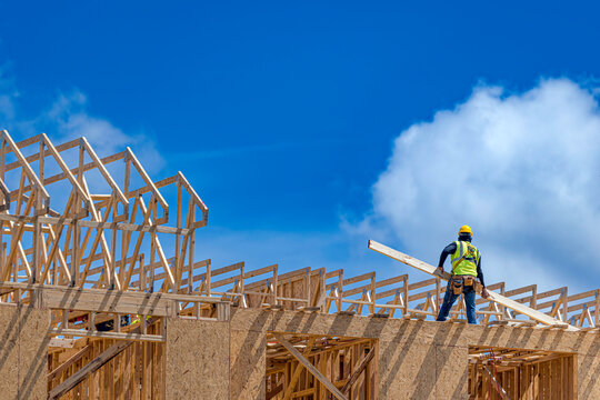 Construction worker standing on the top plate of a wooden house frame and carrying a wood board near roof trusses under a blue sky with clouds, USA