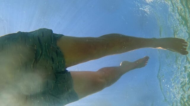 Underwater shot looking up at a man in swim trunks casually swimming by blowing bubbles on a sunny day with blue sky. 