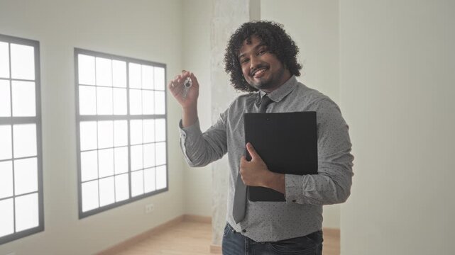 Man holds key in hand and clutches black folder inside empty building room by large window while smiling and presenting keys; confidence new beginning.