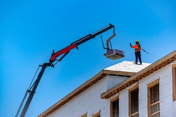 Construction worker standing on a building roof and signaling a hydraulic boom crane lifting a pallet of asphalt shingles roofing materials against a clear blue sky, USA © Andrei Liashchou