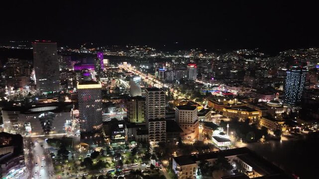 Aerial tracking shot orbiting the illuminated city center of Tirana, showcasing modern skyscrapers, Skanderbeg Square, and vibrant city lights