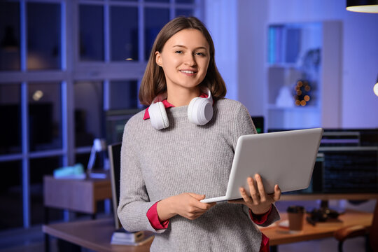 Happy female programmer with laptop and headphones working in office at night
