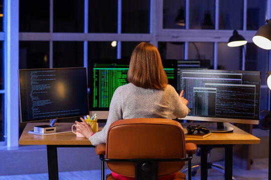 Female programmer with cup of coffee and computers working in office at night, back view
