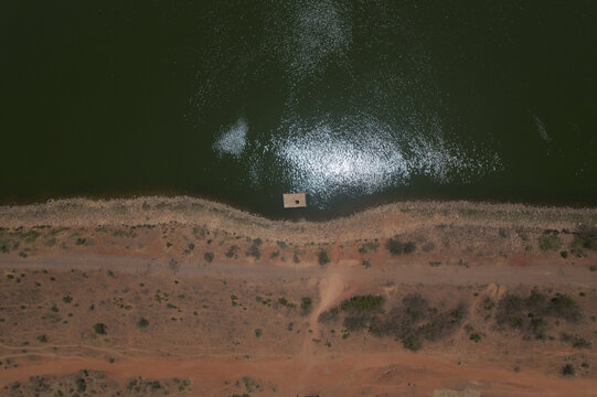 Aerial view of the Minjibir dam with dark green water, sun glint, a small raft near the shore, and an arid landscape with a dirt road in Kano City, Kano State, Nigeria.
